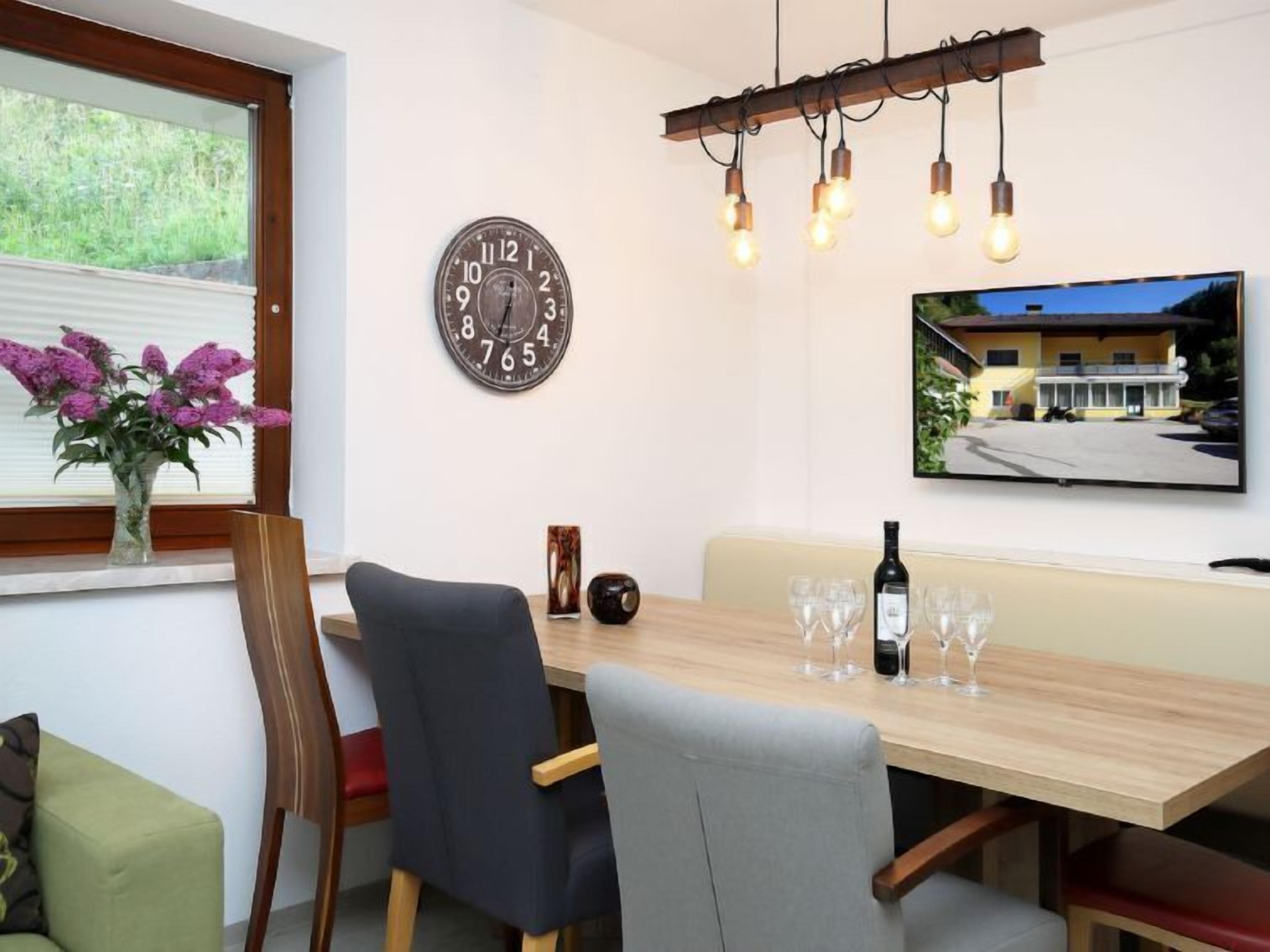 Un comedor con una mesa de madera, sillas, un reloj, un jarrón de flores y un televisor en la pared.