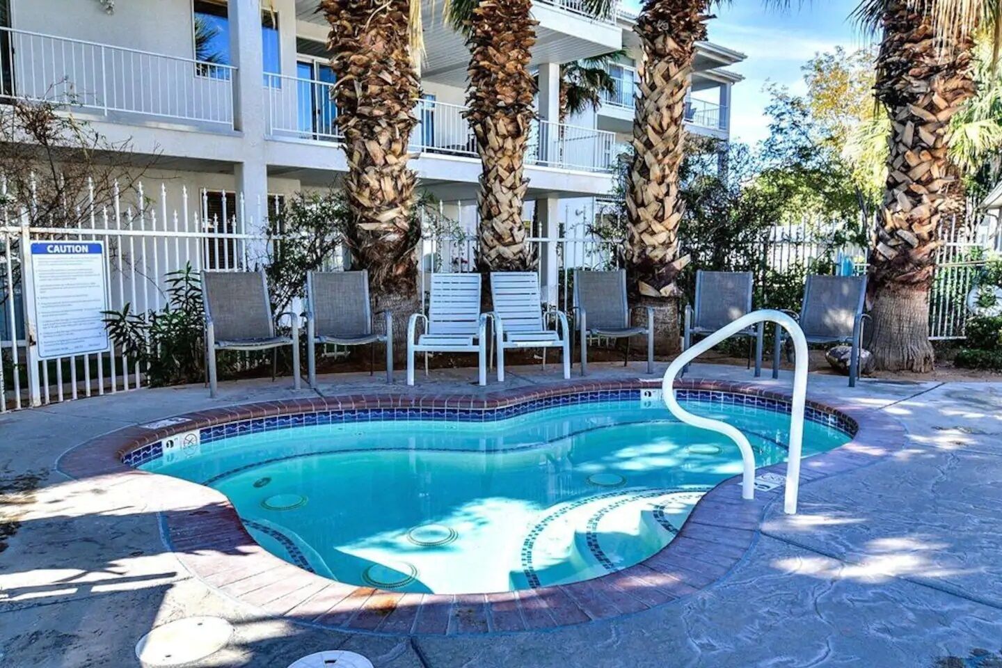 A swimming pool with a tiled edge, surrounded by lounge chairs and palm trees, with a multi-story building in the background.