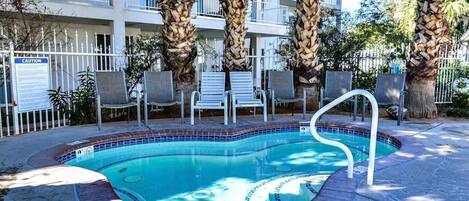 A swimming pool with a tiled edge, surrounded by lounge chairs and palm trees, with a multi-story building in the background.