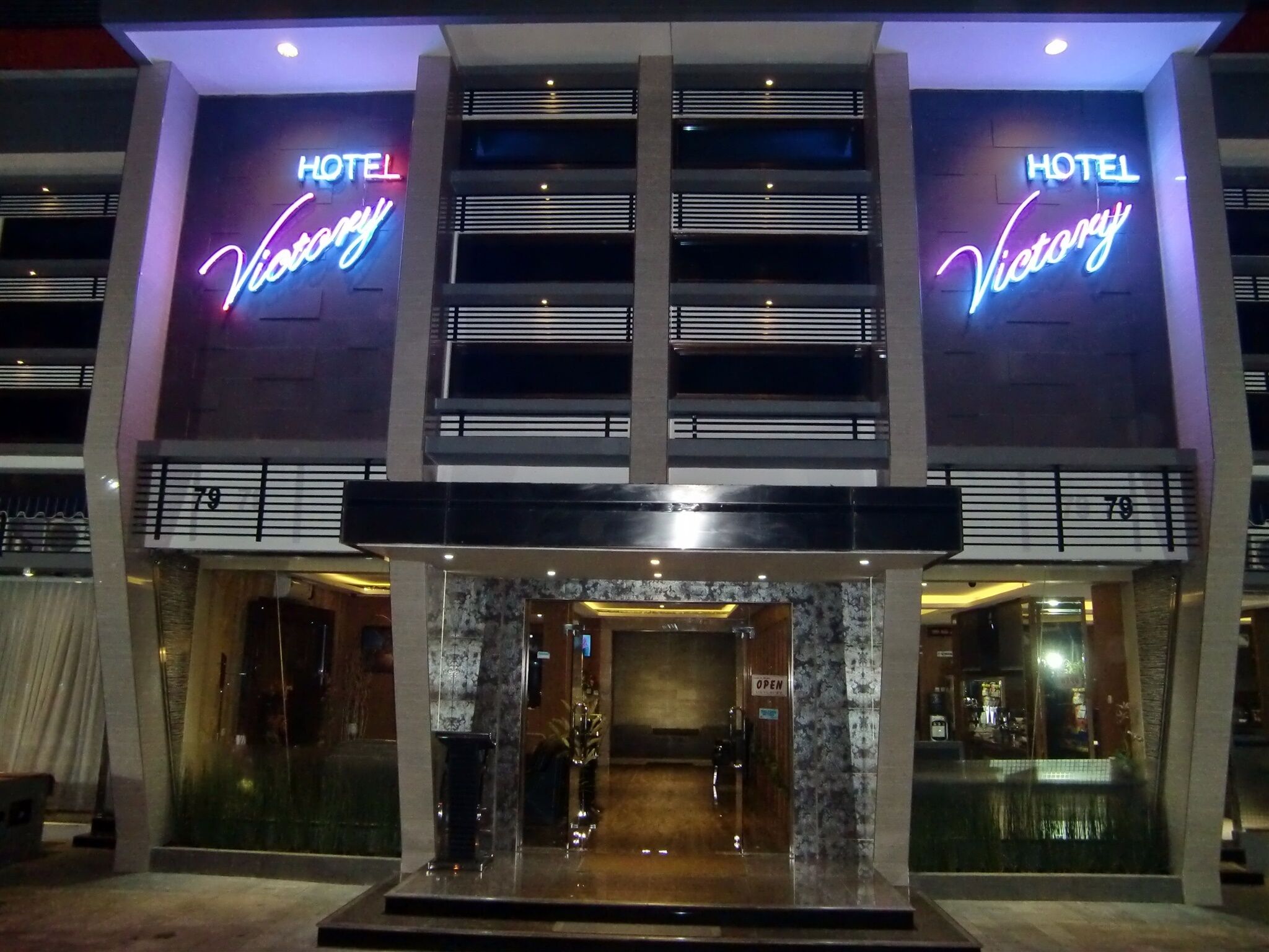 L’entrée d’un hôtel, avec une enseigne au néon indiquant « HÔTEL Victory » et le numéro « 79 » au-dessus de la porte.