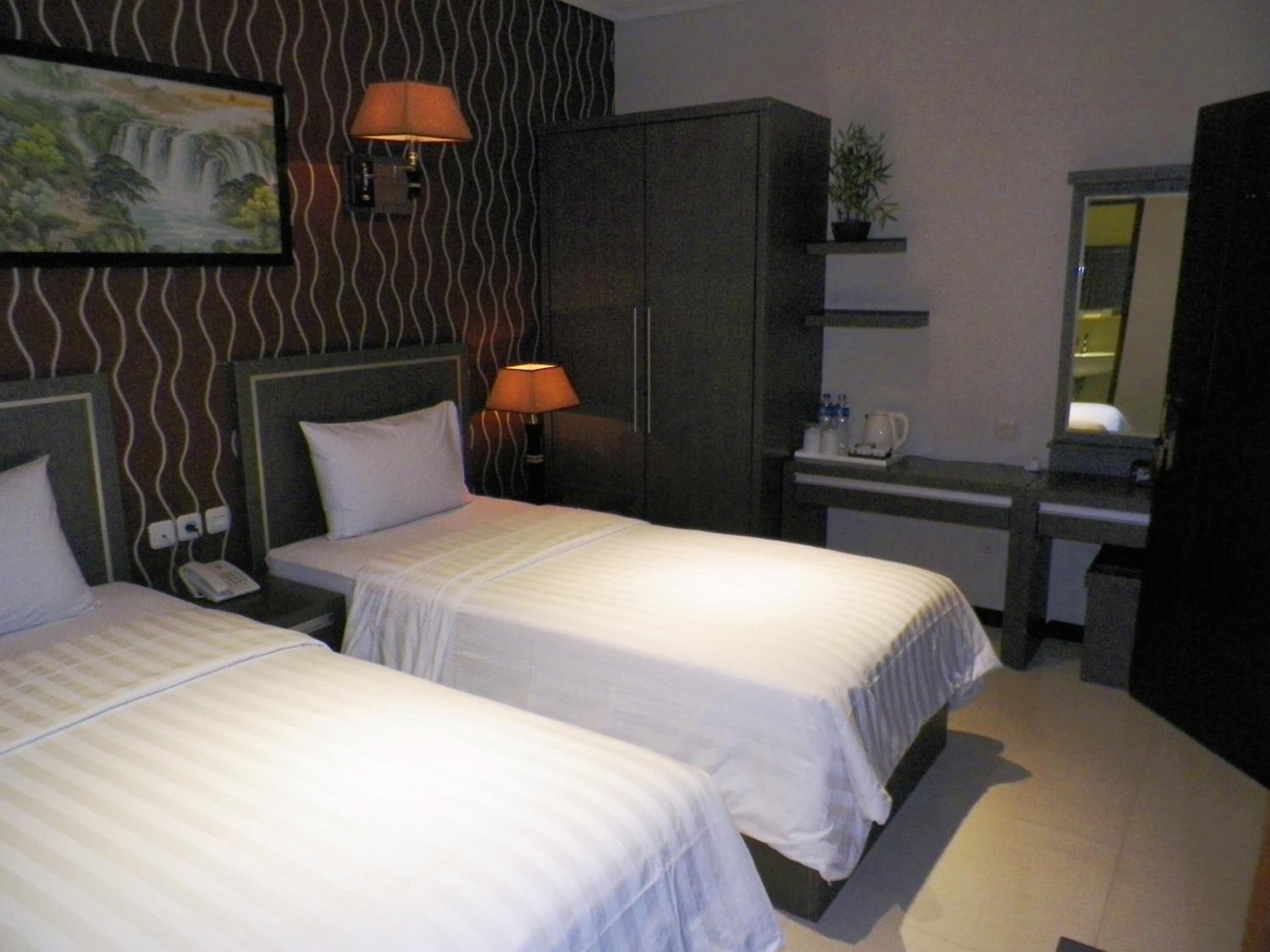 A hotel room with two beds, a desk, a chair, a lamp, a phone, a mirror, and a painting on the wall.