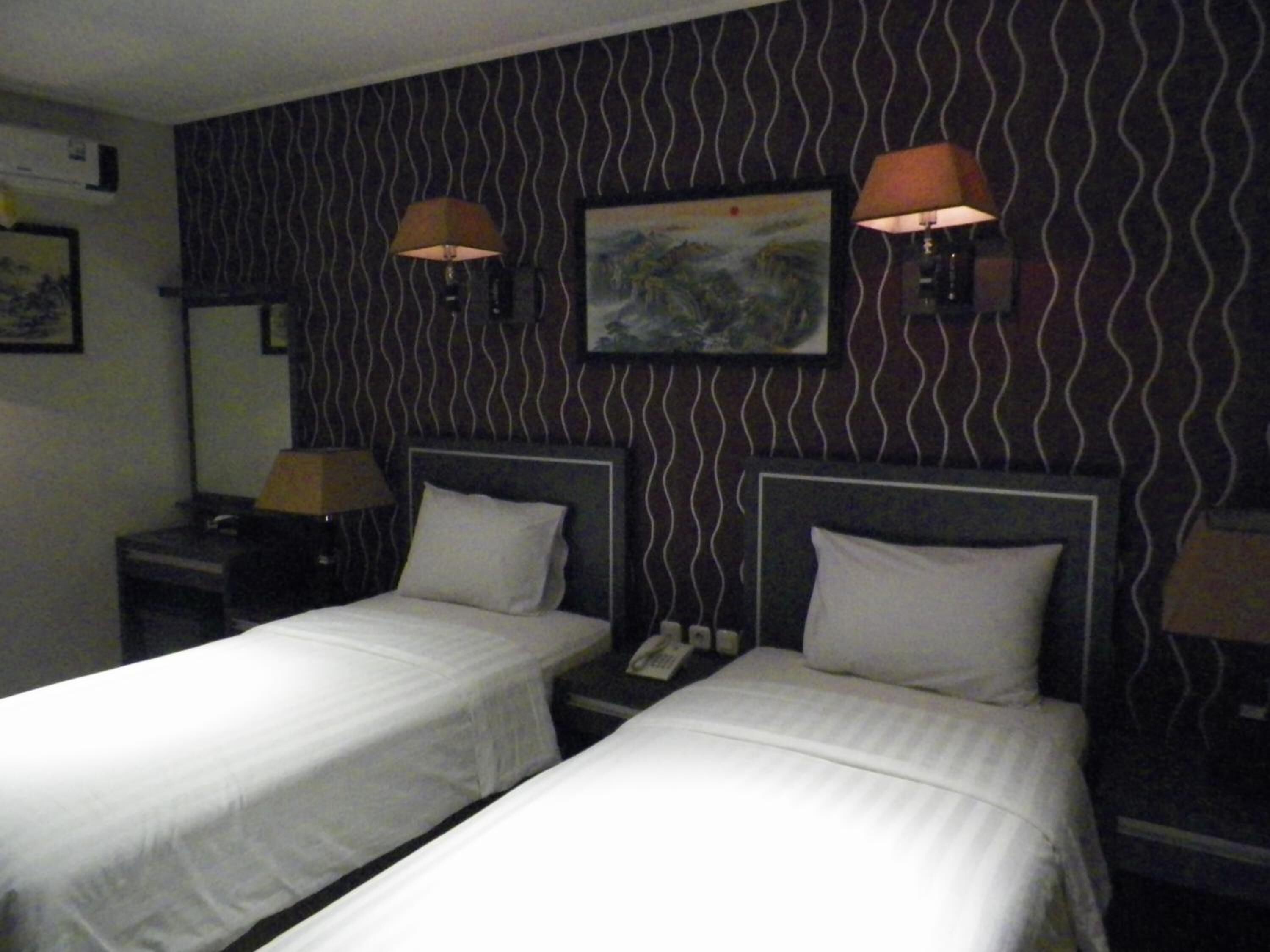 Een hotelkamer met twee eenpersoonsbedden, een nachtkastje met een lamp, een schilderij aan de muur en behang met een patroon.