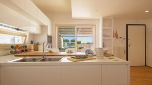 A modern kitchen with white cabinetry, a central island with a sink, and a view through a large window. - TD Villa Fico Jo Villa with Pool & Lush Garden (Conversano)