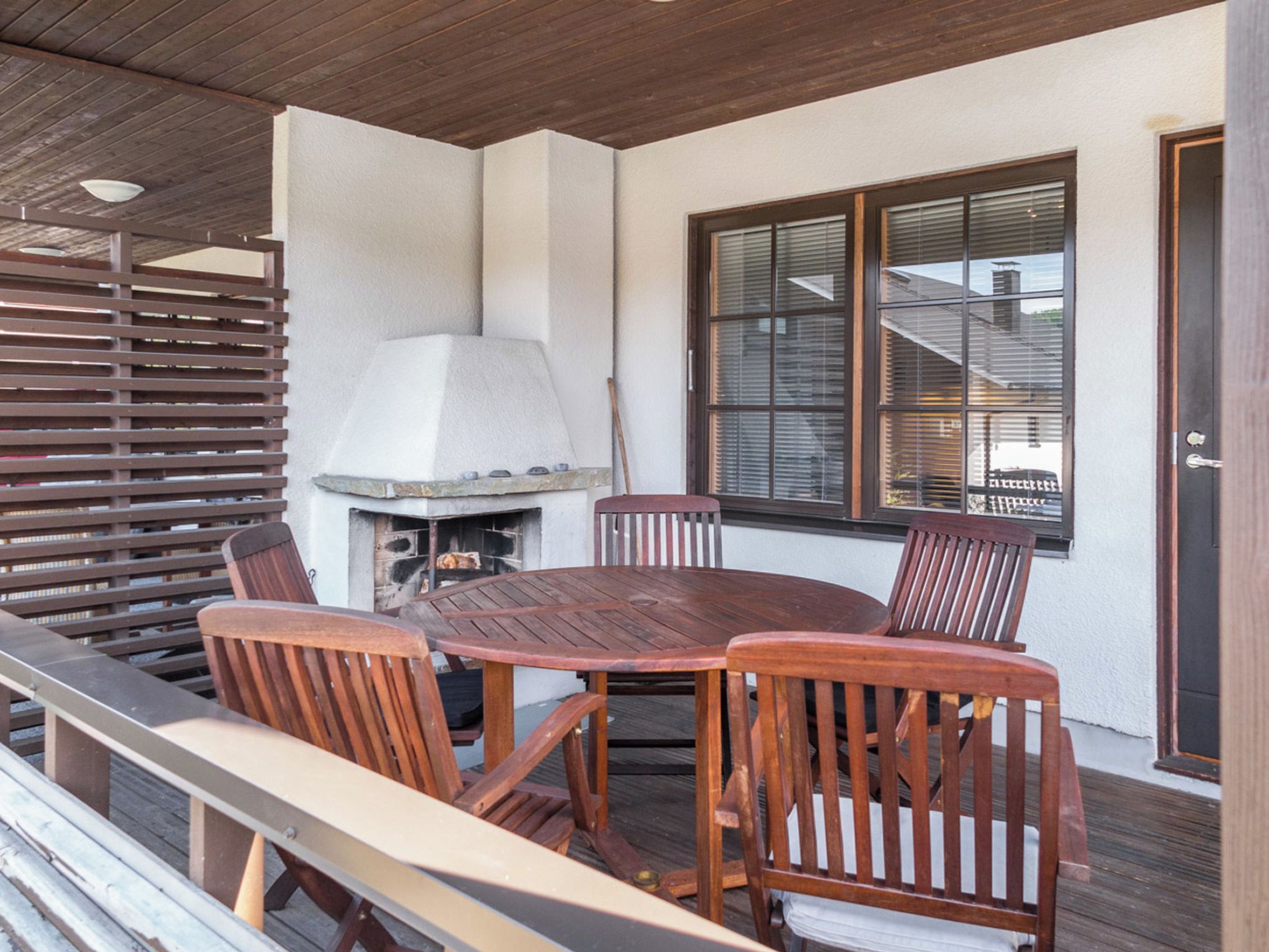 Un patio al aire libre con una mesa redonda de madera y sillas, una parrilla y una ventana con persianas.