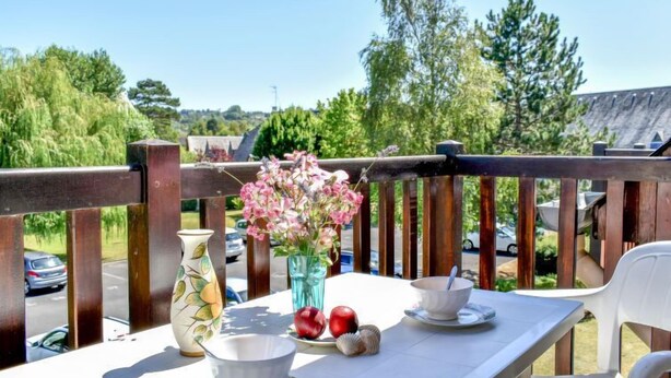 Un balcon avec une table dressée pour deux personnes, un vase rempli de fleurs et une vue sur un parking et des arbres.