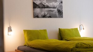 Un dormitorio con cama, dos almohadas, una mesita de noche, un cajón y un cuadro colgado en la pared de un paisaje montañoso.