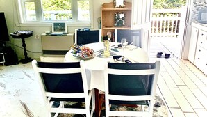 Un coin repas avec une table dressée pour quatre personnes, entourée de chaises. On y trouve une fenêtre avec des stores, une petite table avec une lampe et une étagère ornée d’objets décoratifs.