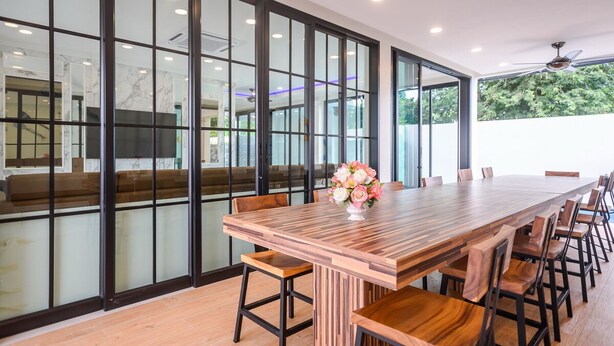 A long wooden conference table with matching chairs, a vase of flowers, and a modern interior with large glass doors and a ceiling fan. - Pool Villa Pattaya - Exquisite Pool Villa H (Pattaya City)