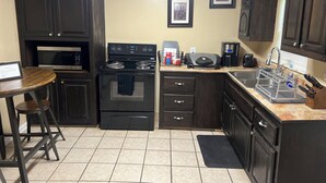 A compact kitchen with dark cabinetry, a microwave, stove, and a coffee maker. There is a bar stool and a small table with a framed certificate. - My Lil Red House.... (Baton Rouge)