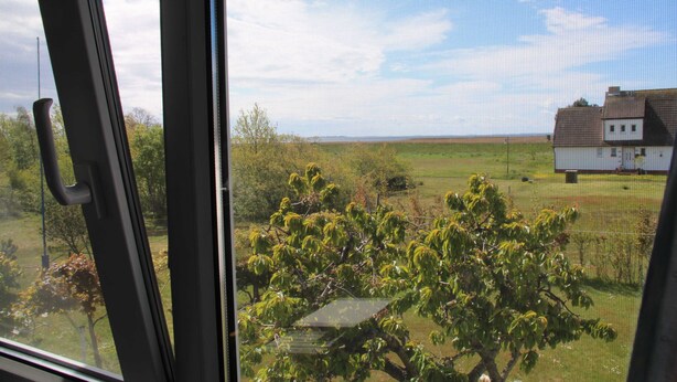 Vista pela janela de uma paisagem rural com uma casa e campos abertos.