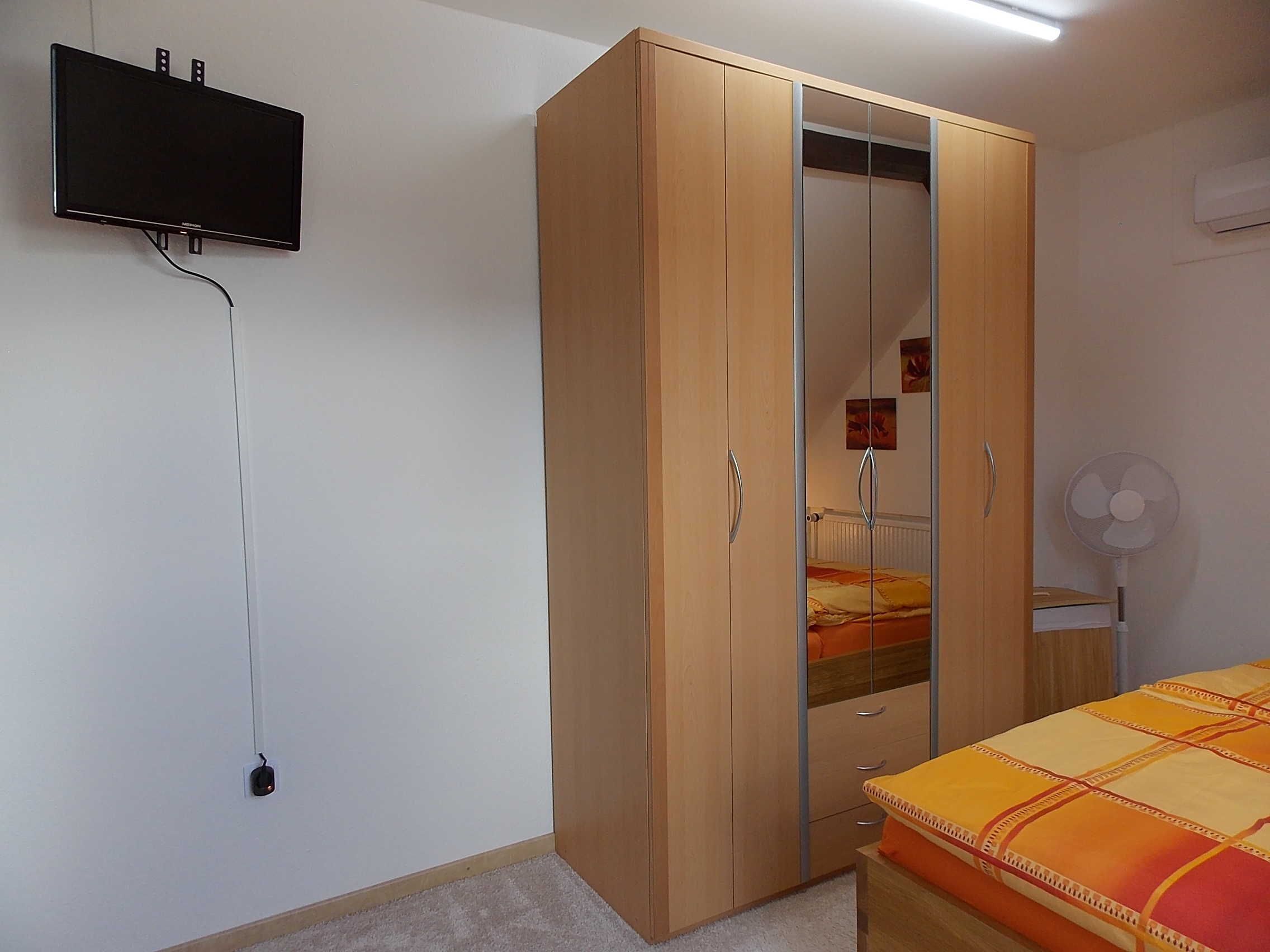 A bedroom with a wardrobe, a bed with a patterned bedspread, a wall-mounted TV, and a standing fan.