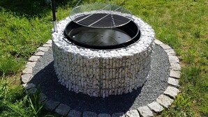 A circular stone fire pit with a metal grill, surrounded by a stone border, in a grassy outdoor area with a wooden bench and trees in the background. - Ferienwohnung Mohnblume (Gerswalde)