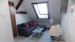 A small, modern room with a dark grey sofa, a white coffee table, and a glass lamp. There is a skylight window above the sofa and a white fan on the wall. - Ferienwohnung Mohnblume (Gerswalde)
