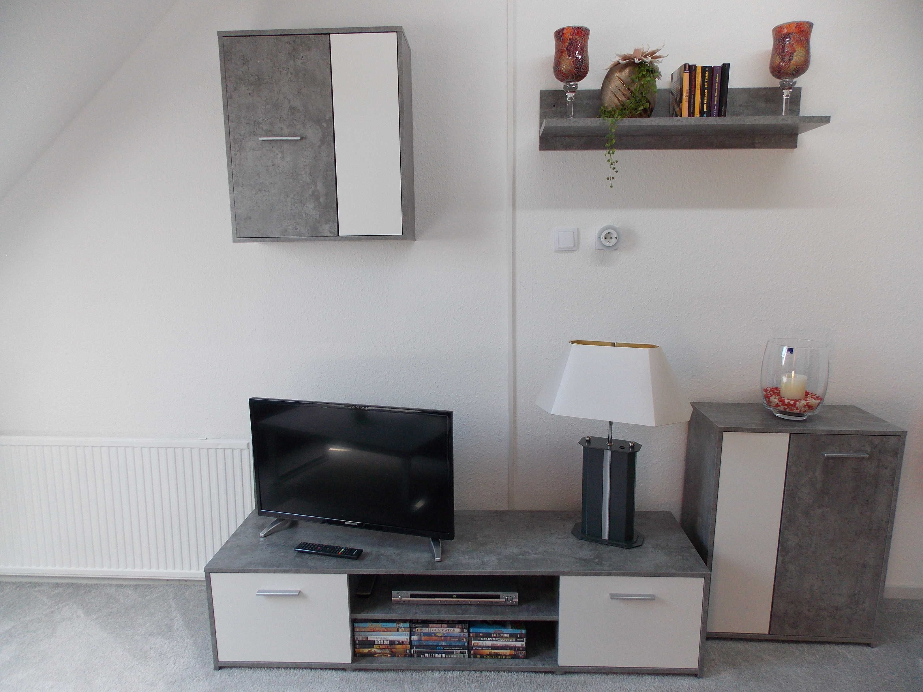 A modern living room with a flat-screen TV on a grey entertainment unit, a lamp, and a small shelf with decorative items.