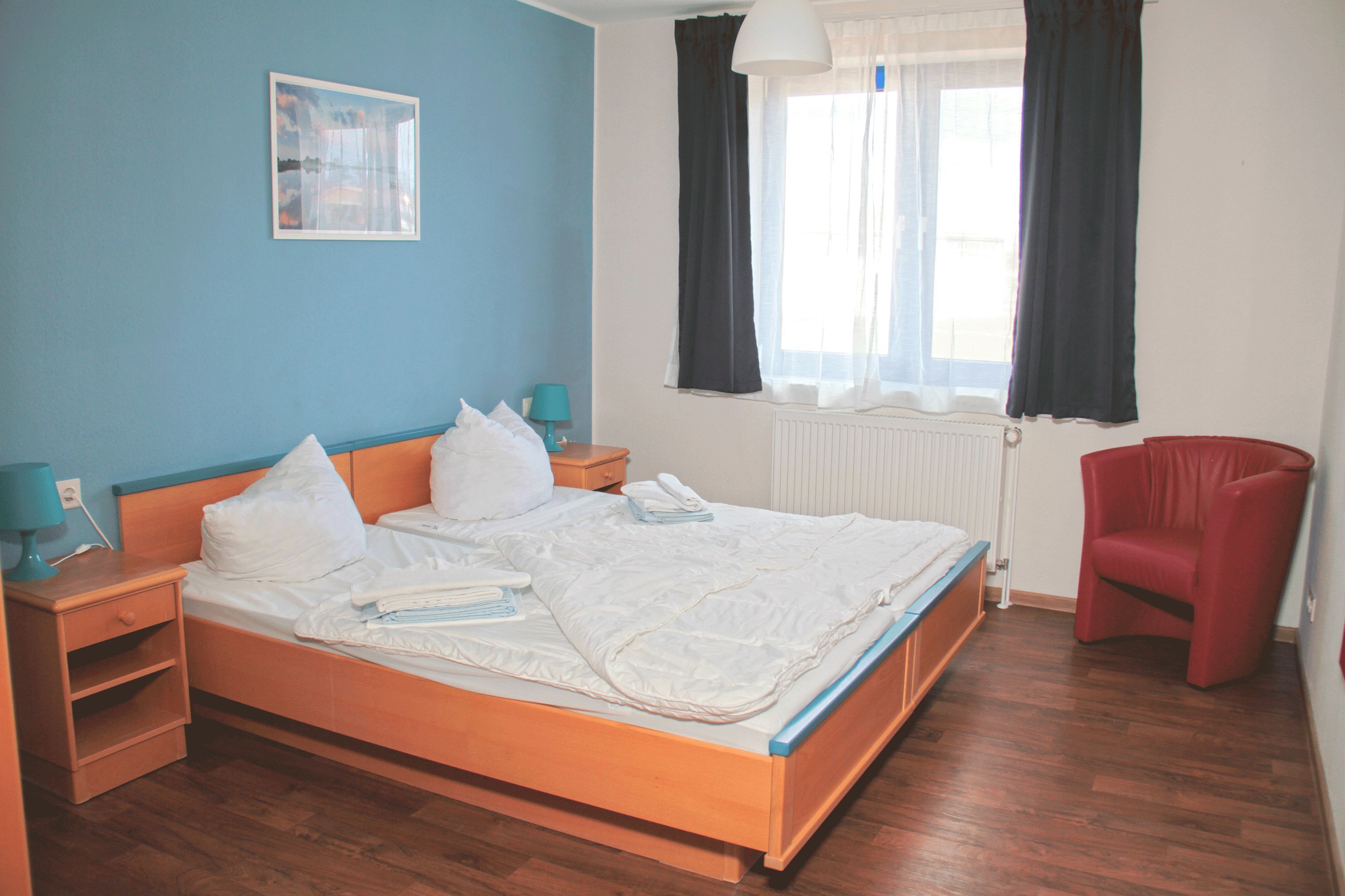 Ein Schlafzimmer mit einem hölzernen Bett, einem Nachttisch, einem roten Sessel, einem Fenster mit schwarzen Vorhängen und einem an der Wand hängenden Bild.