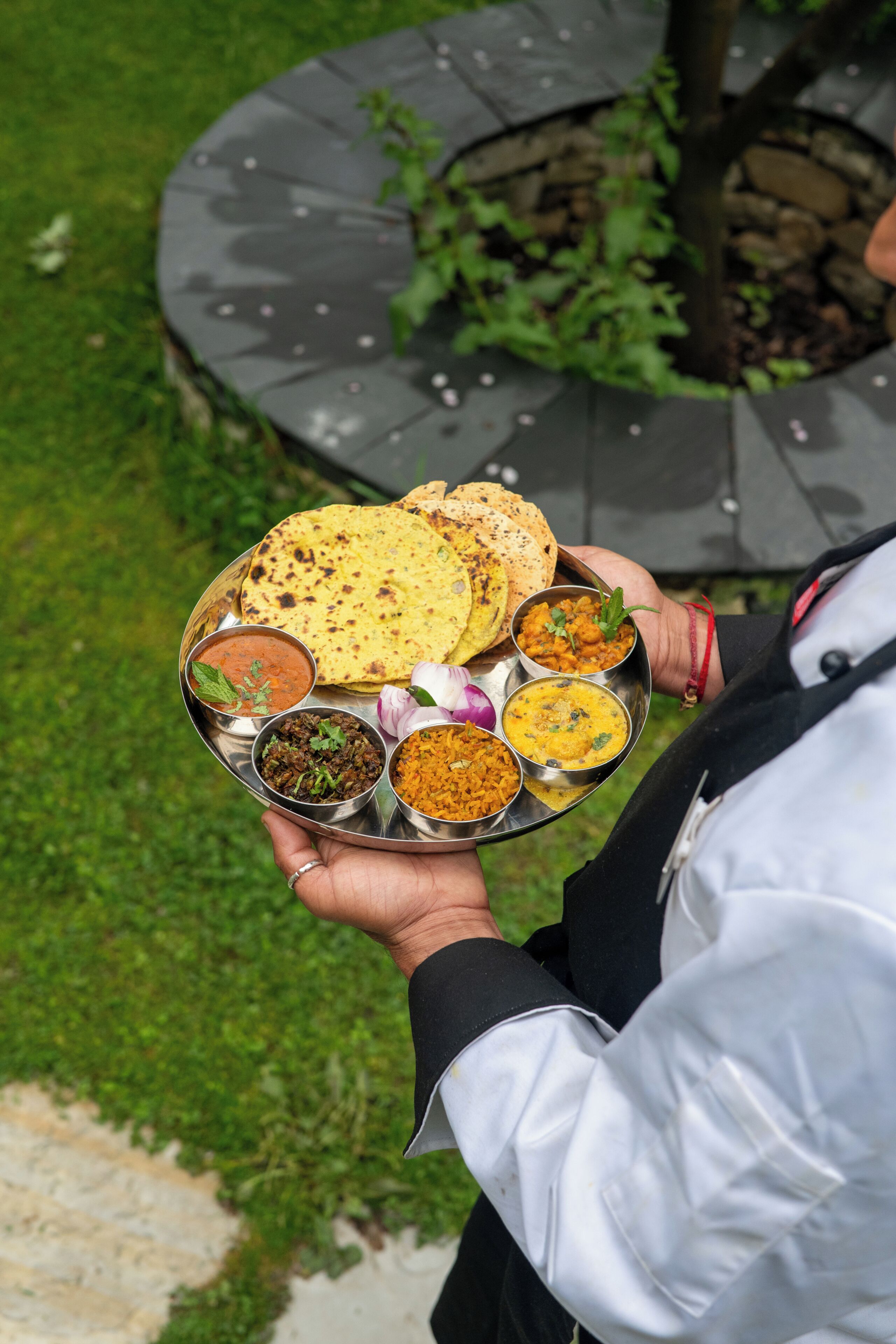 A person holding a metal tray with various Indian dishes.