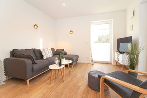 A modern living room with a grey sofa, a small round table, a TV on a stand, and a large window.