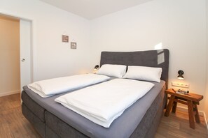 A neatly made bed with white linens, a grey upholstered headboard, and a small bedside table with a lamp.