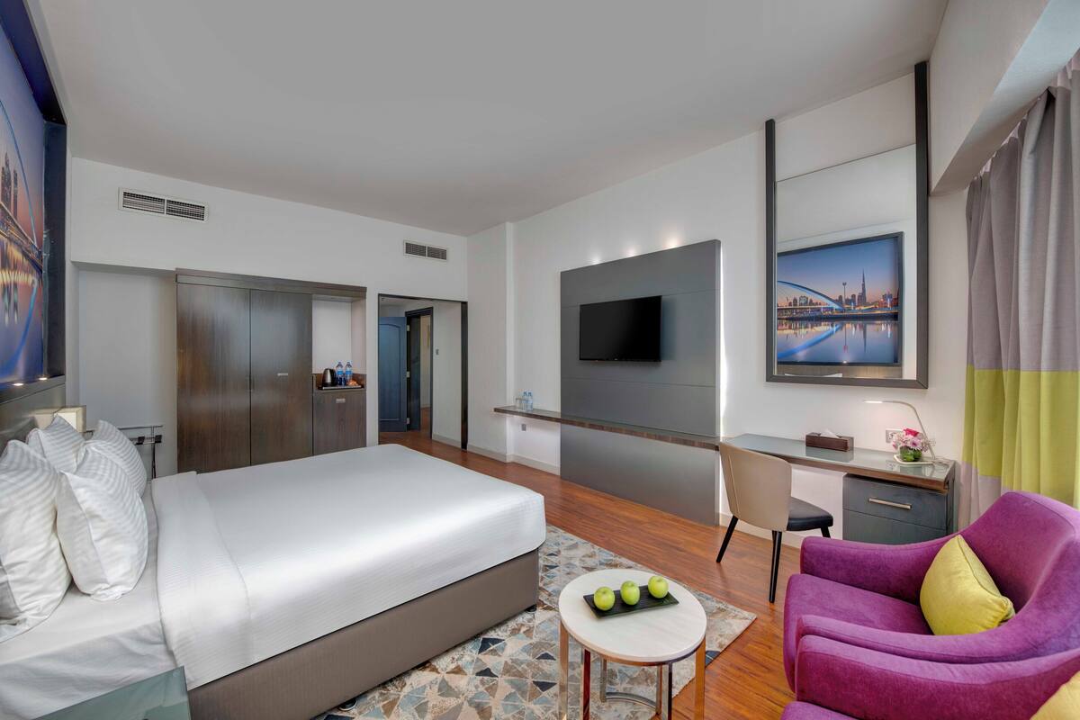 A modern hotel room with a large bed, a desk with a computer, a TV, and a cityscape painting.
