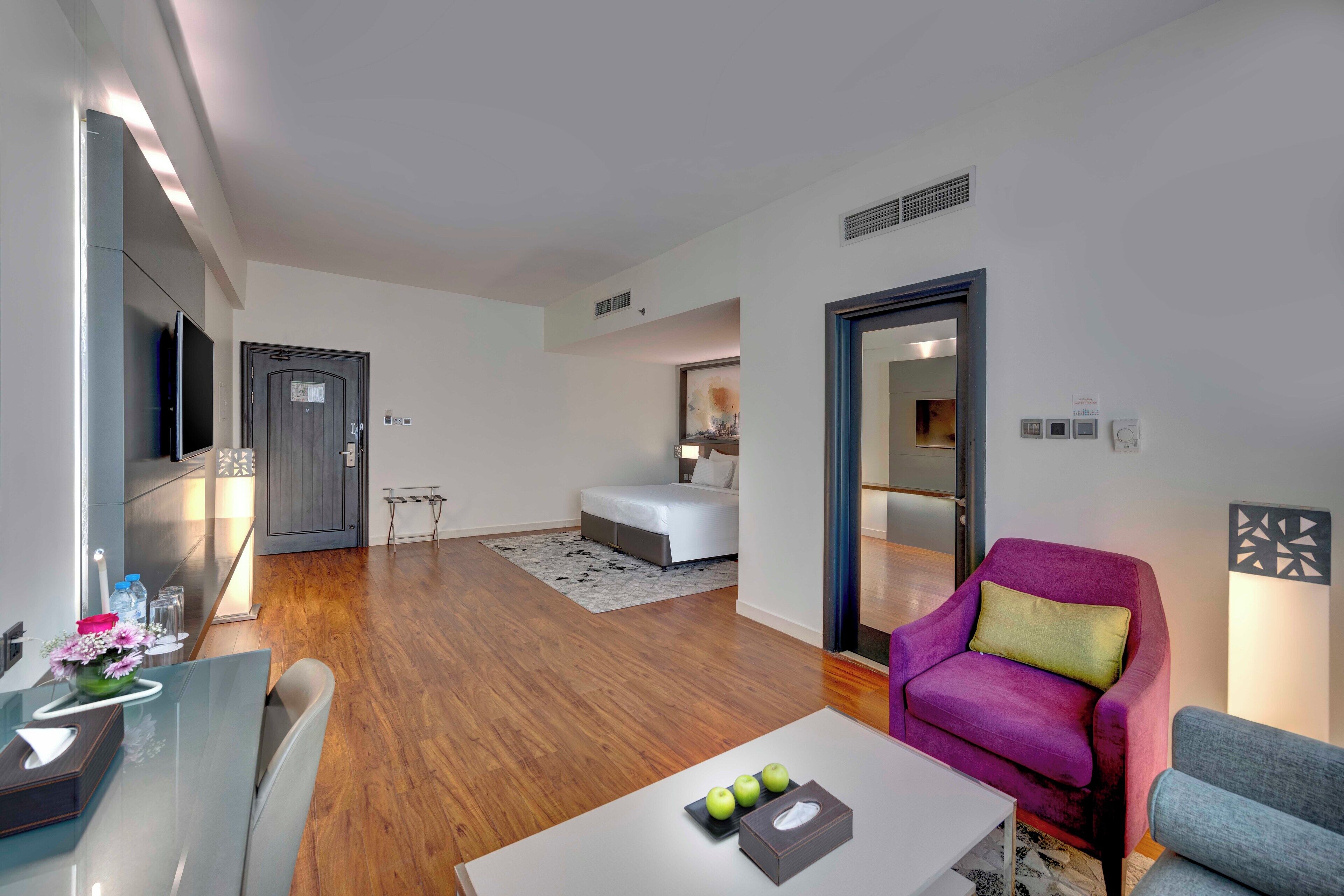 Un salón moderno con una mesa de comedor de vidrio, un sillón morado y un sofá blanco. Hay una mesita auxiliar con un jarrón de flores y un control remoto.