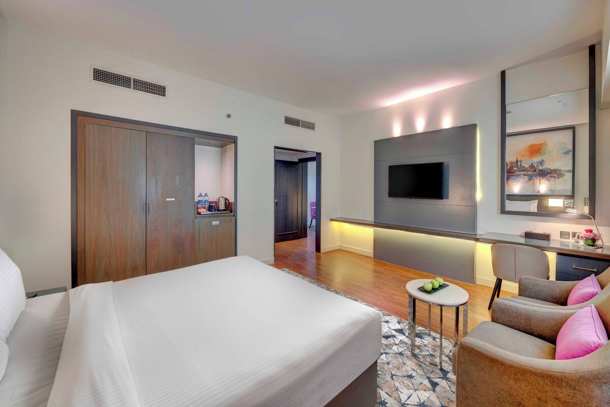 A modern hotel room with a large bed, a seating area with a small table, a TV mounted on the wall, and a kitchenette.