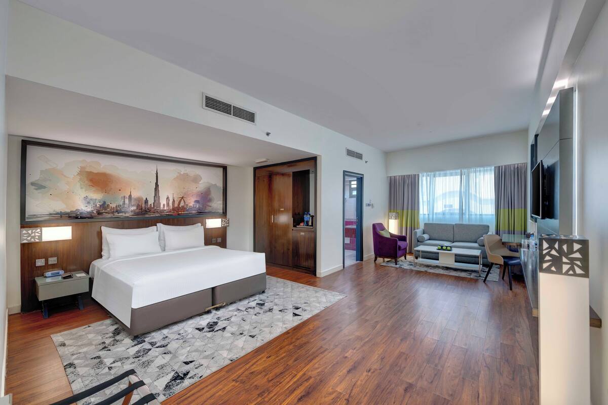 A modern hotel room with a large bed, a desk, a sofa, and a cityscape painting on the wall.