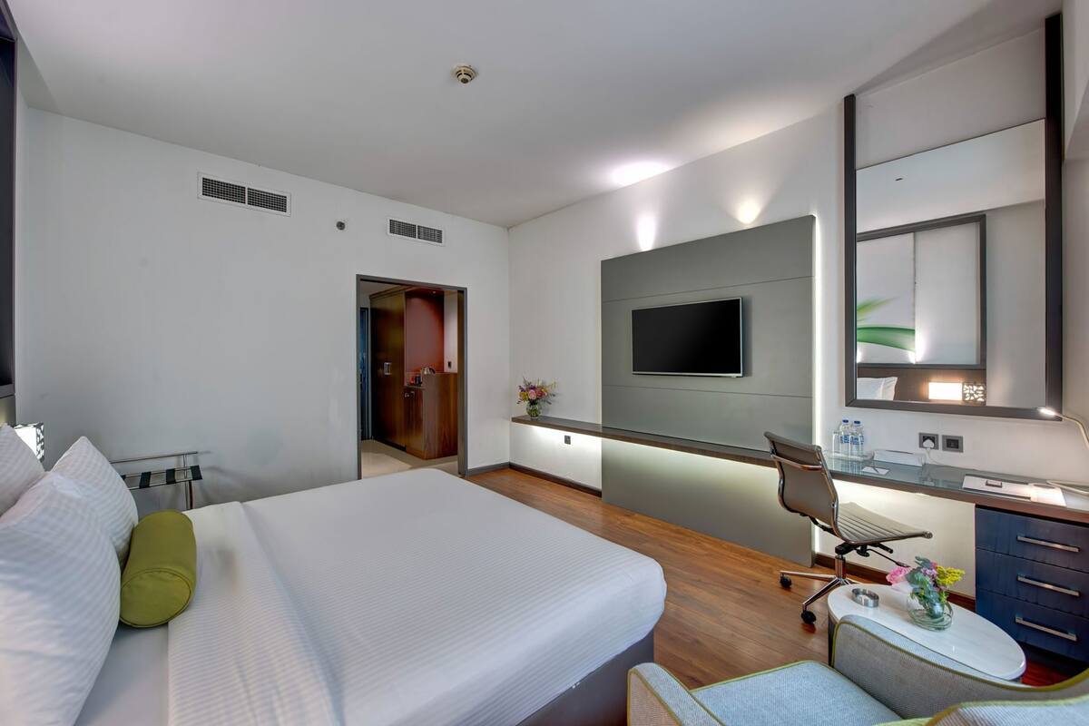 A hotel room with a bed, a desk with a chair, a TV, a mirror, and a small table with a vase of flowers.
