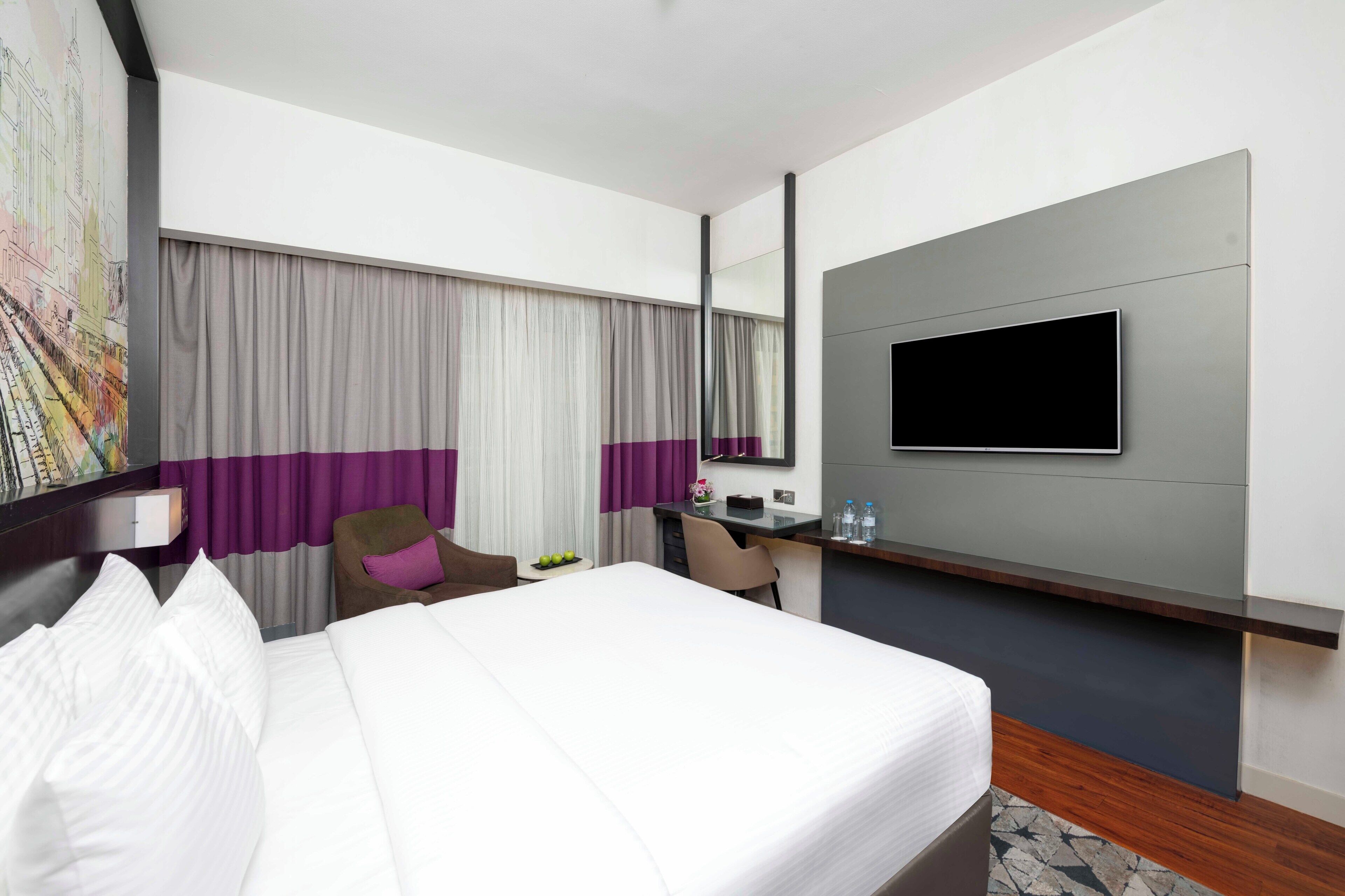 Une chambre d’hôtel moderne dotée d’un grand lit, d’un bureau, d’une télévision à écran plat et d’une grande fenêtre avec des rideaux.