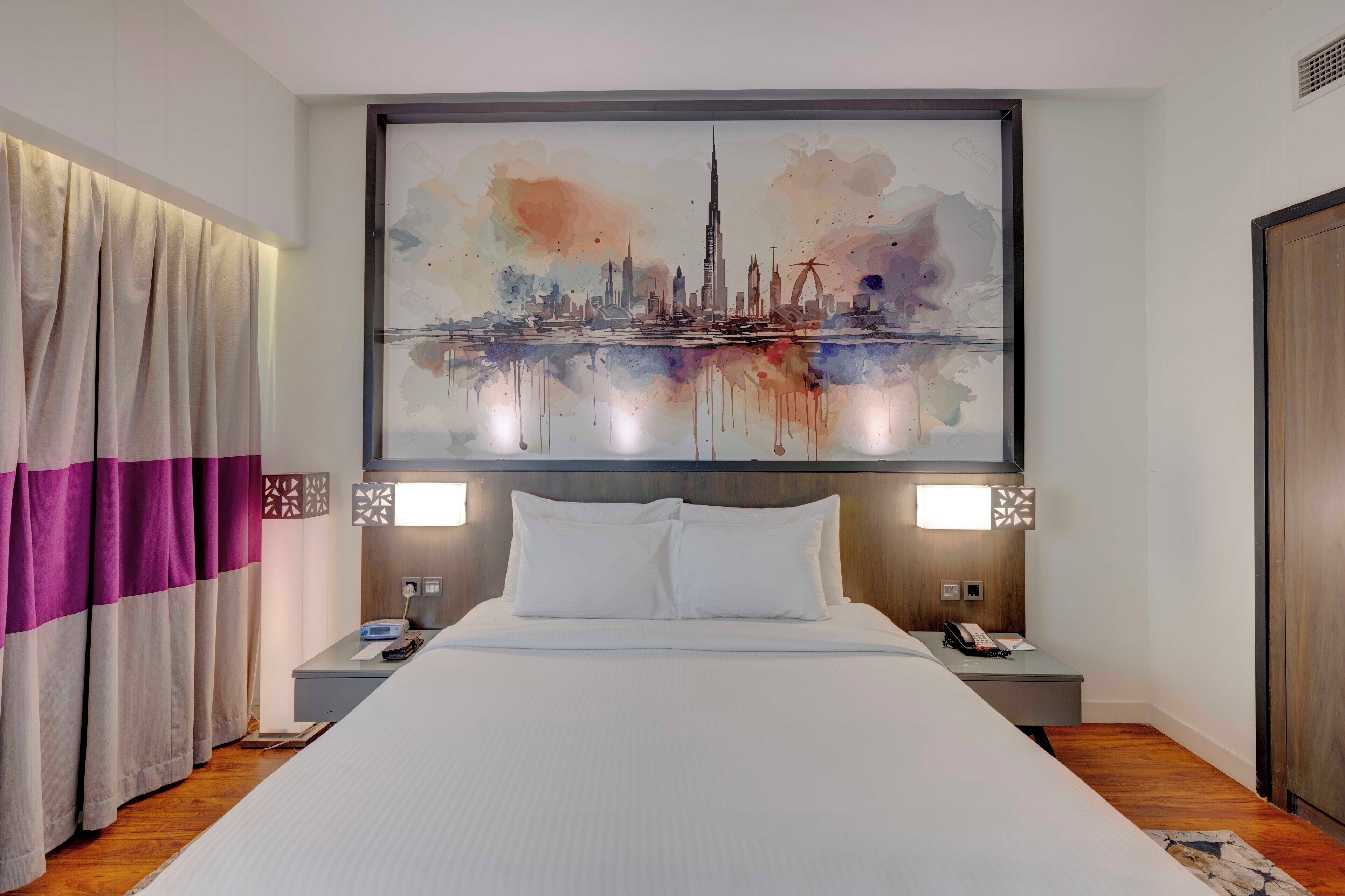 Ein Hotelzimmer mit einem großen Bett, zwei Nachttischen, einem Stadtbild an der Wand und einem Vorhang mit violetten und weißen Streifen.
