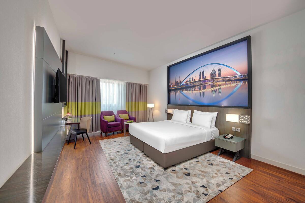 A modern hotel room with a large bed, a flat-screen TV mounted on the wall, a cityscape mural, a dining area with a table and chairs, and a small side table with a lamp.
