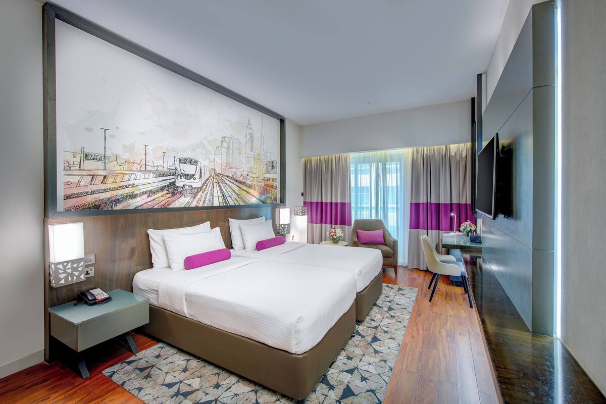 A modern hotel room with a large bed, a desk, a chair, a TV, and a cityscape painting on the wall.