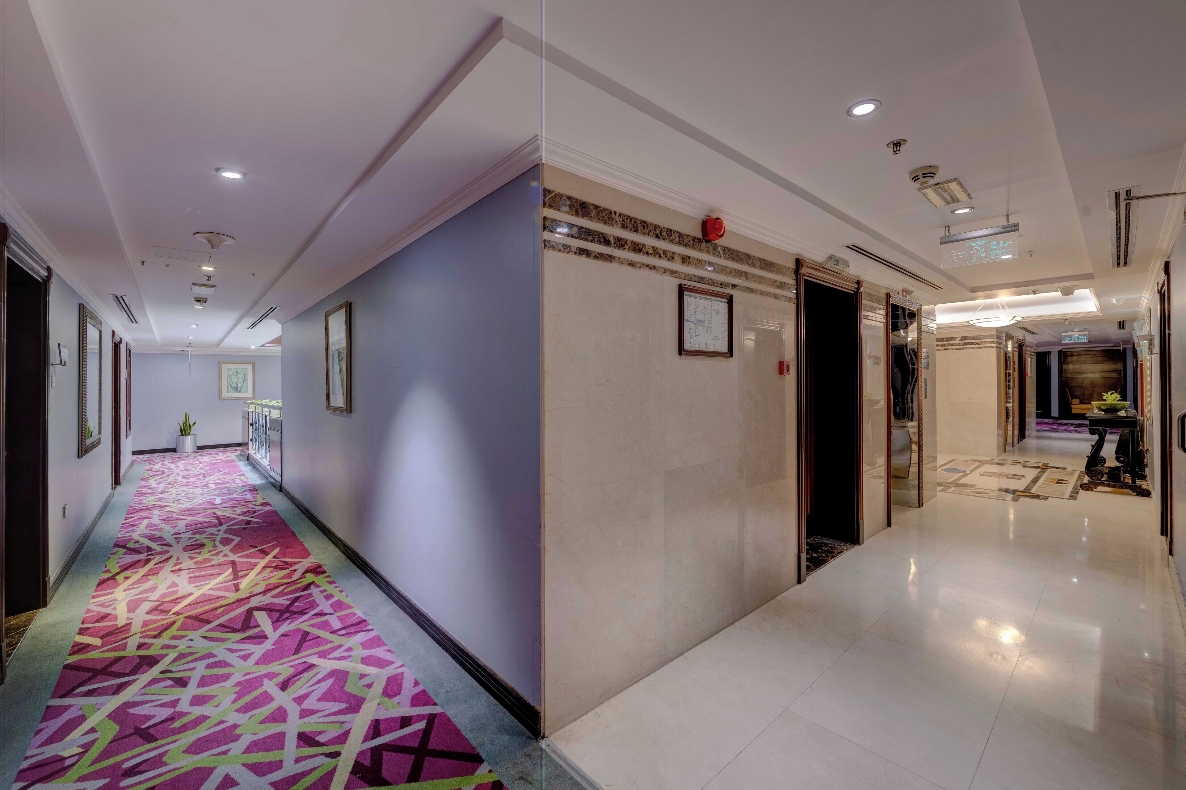 A hotel corridor with a patterned carpet, framed artwork on the walls, and a clean, tiled floor.