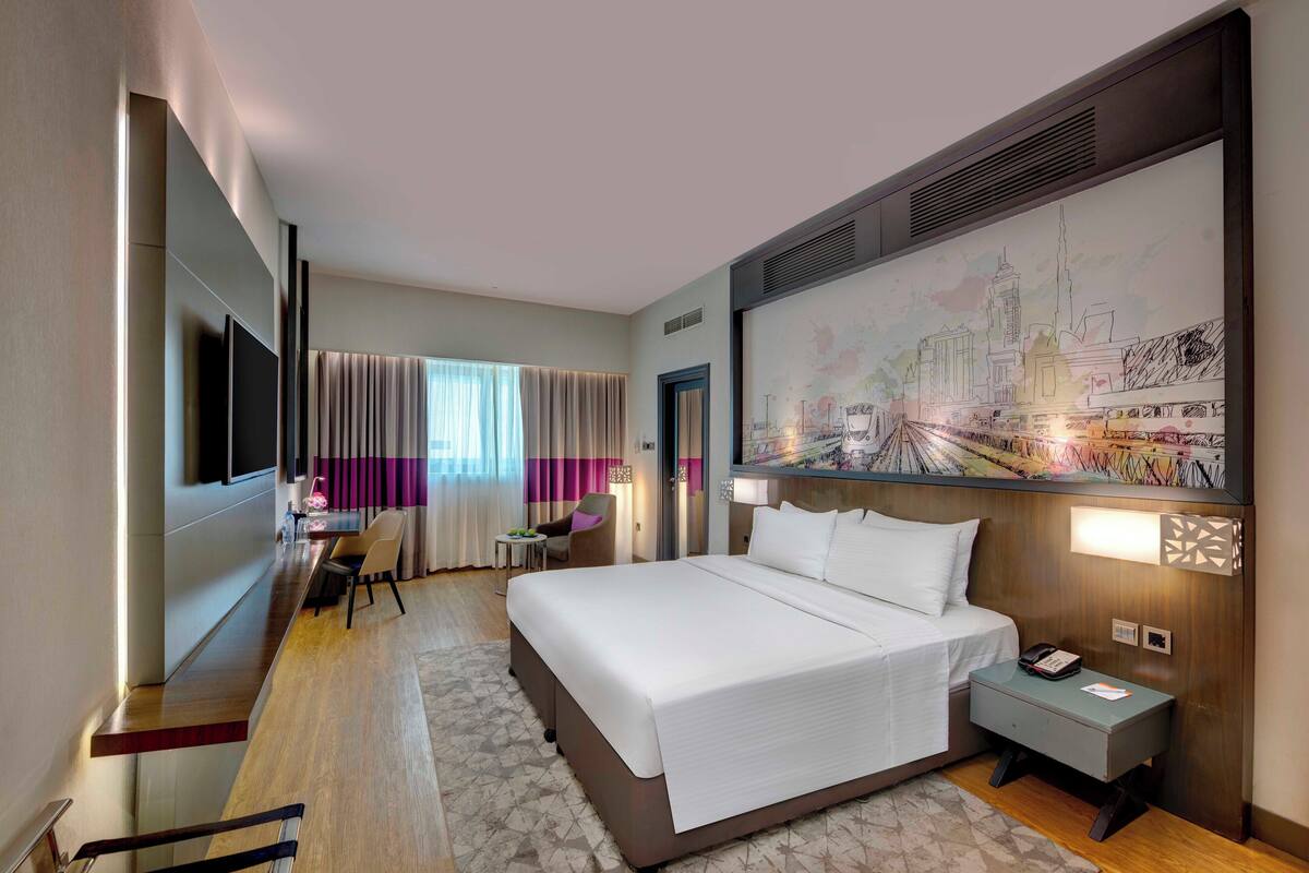A modern hotel room with a large bed, a desk, and a chair. There is a painting on the wall and a window with curtains.