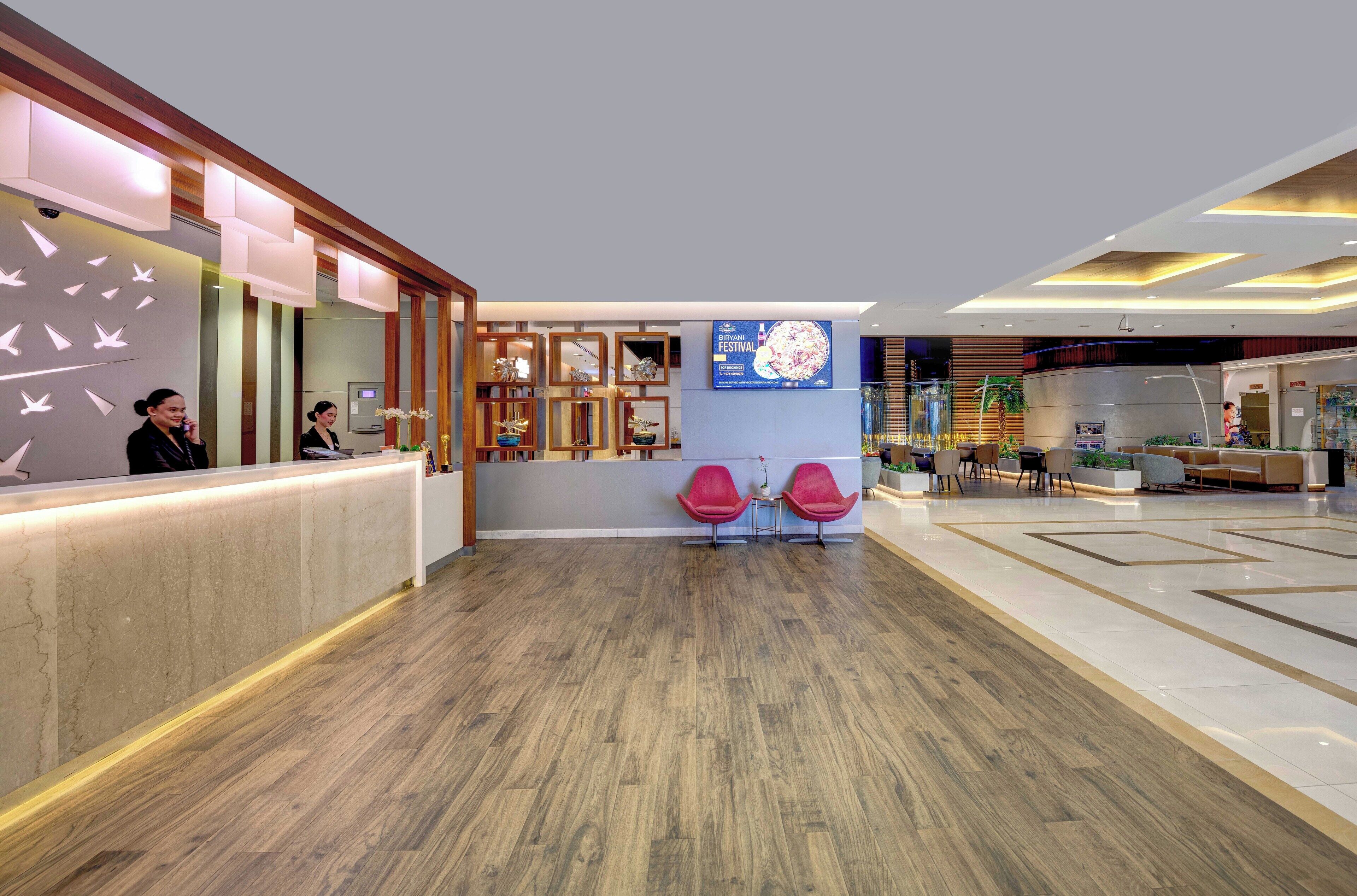 A modern hotel lobby with a reception desk, wooden flooring, and a large digital screen displaying a menu.
