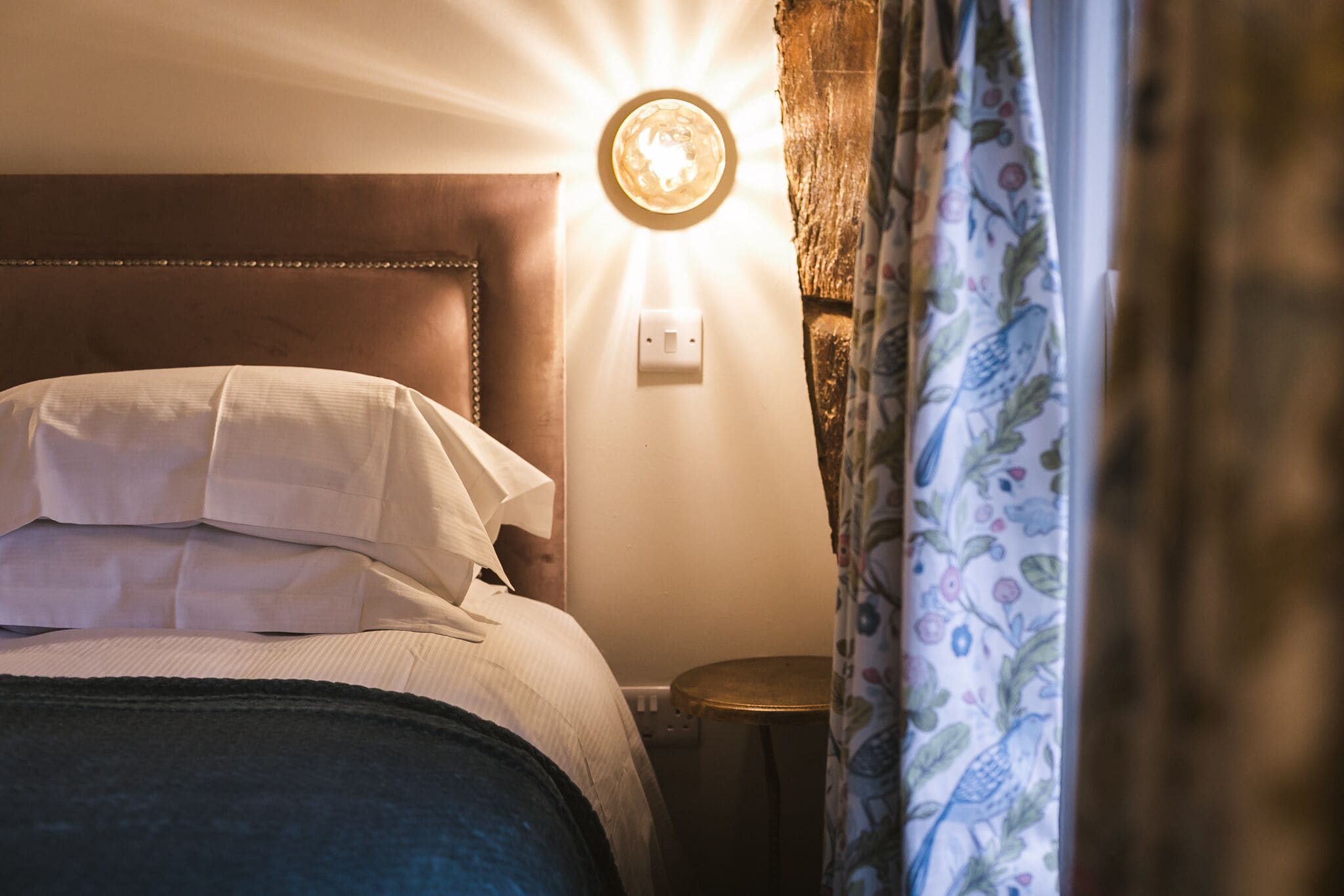 A bed with white linens, a round wall light, and a small round table beside it.
