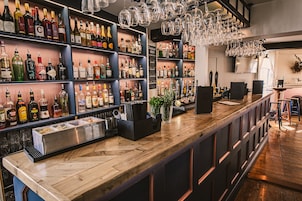 Una zona de bar con estantes llenos de botellas de licores, una barra de madera y copas colgando.