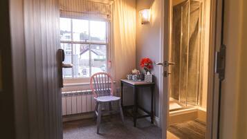 Ein Badezimmer mit Dusche, einem fenster mit Vorhängen, einem Heizkörper, einem Stuhl und einem kleinen Tisch mit Blumen.