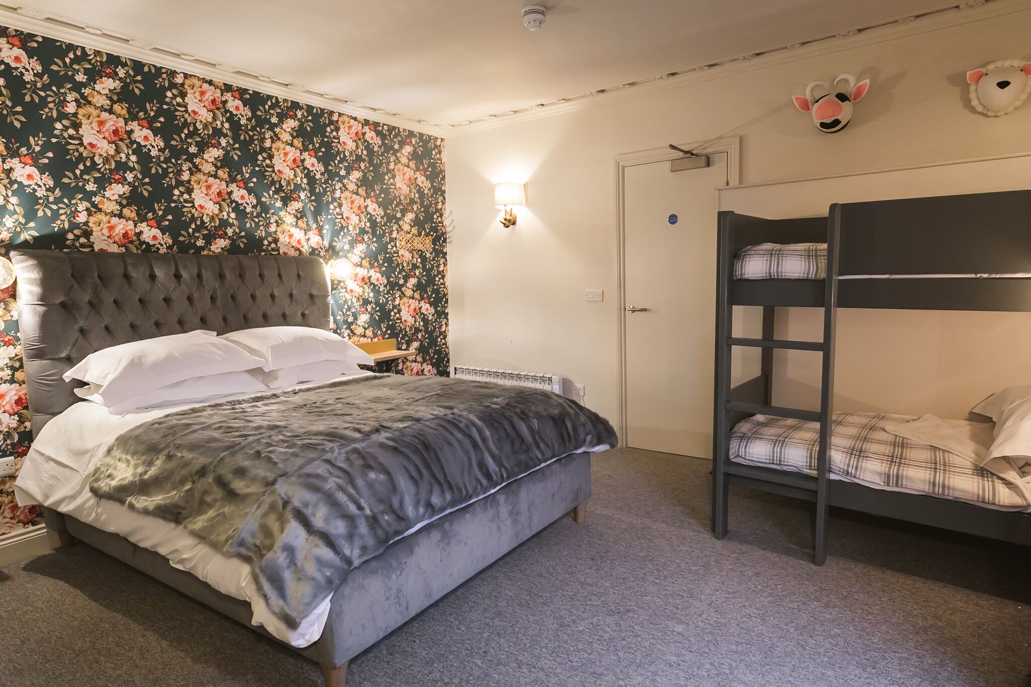 A bedroom with a tufted headboard bed, a bunk bed, floral wallpaper, and a wall-mounted lamp.