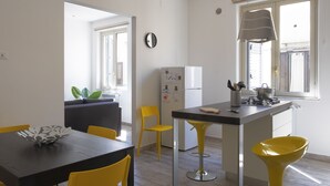 Una cocina moderna con zona de comedor, sillas amarillas, una mesa negra y un ventanal con persianas.
