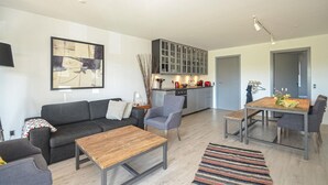 A modern living room with a grey sofa, a wooden coffee table, and a dining area with a wooden table and chairs. There is a kitchen with grey cabinets and a hanging light fixture.