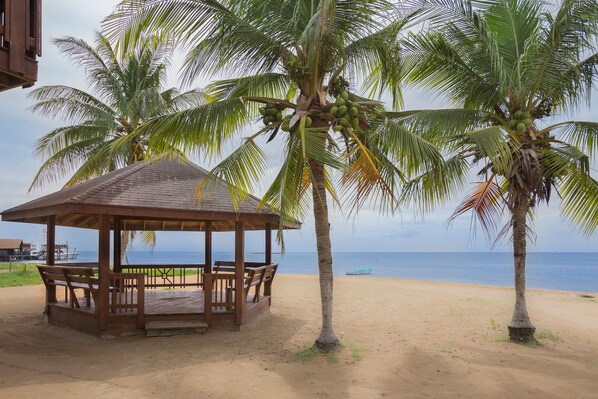 Un quiosco de madera con bancos sobre una playa arenosa, palmeras y el océano al fondo.