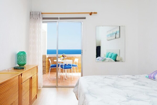 Une chambre avec un lit, une commode en bois, un vase vert, un balcon avec une table et des chaises, et une vue sur la mer.