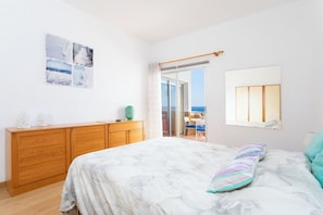 A bedroom with a bed, a wooden dresser, a window with a view, and a wall-mounted lamp.