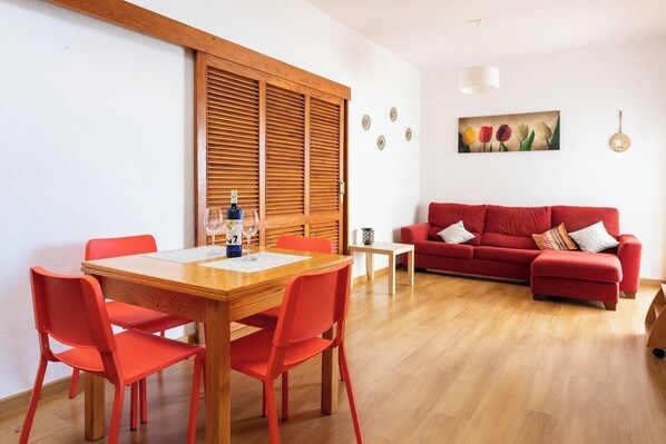 A living room with a red sofa, a wooden dining table with four chairs, and a window with wooden shutters.