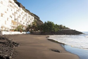 A coastal hotel with white balconies, a sandy beach, and a rocky cliff.