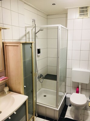 Une salle de bain avec une douche à parois de verre, un lavabo blanc, des toilettes et une petite étagère contenant des articles de toilette.