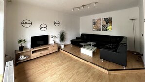 A modern living room with a flat-screen TV, a black sofa, a coffee table, and two potted plants. There are three wall-mounted light fixtures and two framed artworks. - Ferienwohnung Ursulastraße (Dinslaken)