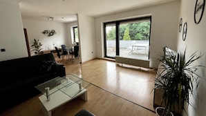 A spacious living room with a large sliding glass door leading to a balcony, a black sofa, a glass coffee table, and a potted plant. - Ferienwohnung Ursulastraße (Dinslaken)