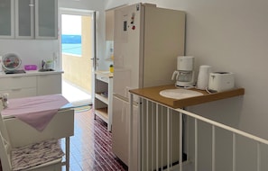 A compact kitchen with a refrigerator, coffee maker, and toaster. A small dining table with a chair is visible, and a window opens to a balcony. - Amazing apartment in Dugi Rat with house sea view (Dugi Rat)