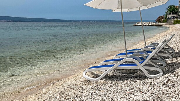 Ein Strand mit klarem Wasser, eine Reihe Sonnenliegen und ein weißer Sonnenschirm.